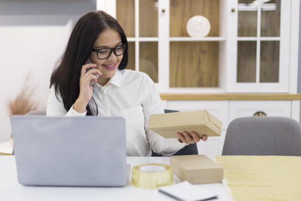 Impiegata con occhiali e camicia bianca sorride mentre parla al telefono, osservando un pacco sulla scrivania accanto a un laptop e materiali per la spedizione.