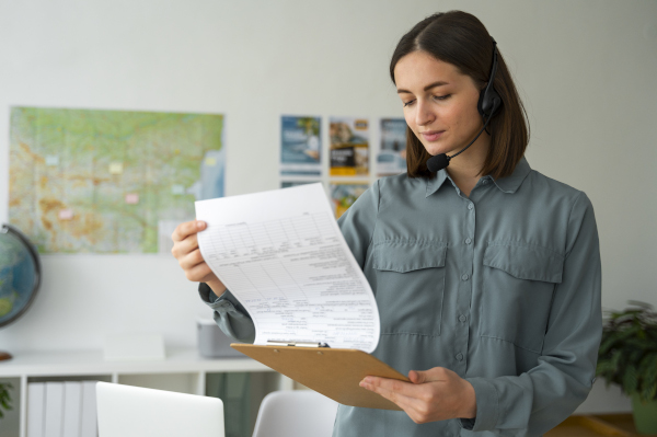 Impiegata logistica con camicia verde e cuffia con microfono analizza un documento su una cartellina, seduta a una scrivania con mappe e documenti di logistica.