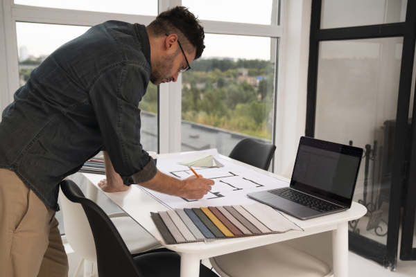 Uomo interior designer lavora a una planimetria su un tavolo con un computer e campioni di tessuti, in una stanza luminosa con grandi finestre.